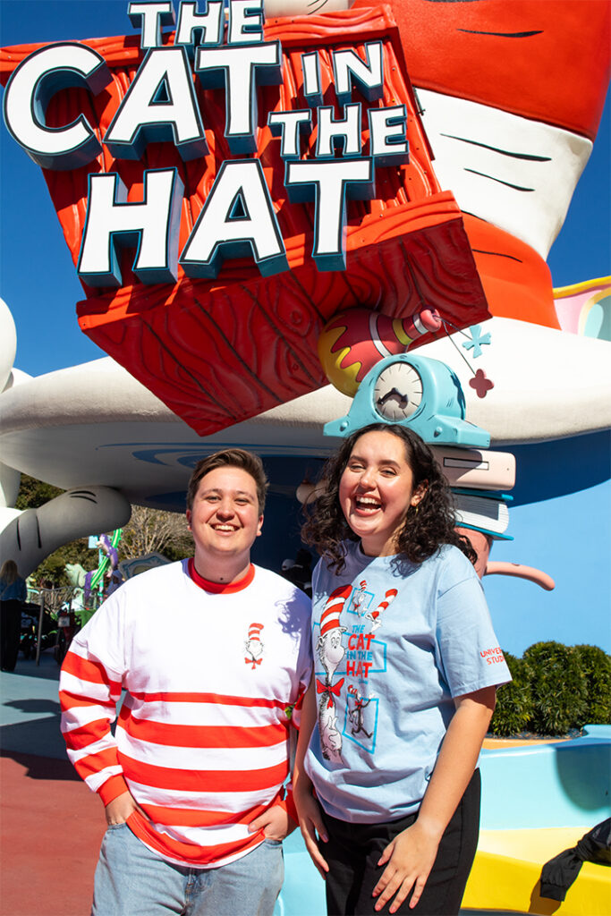 Two friends smile and pose in Cat in the Hat apparel in front of the attraction in Seuss Landing at Universal Islands of Adventure