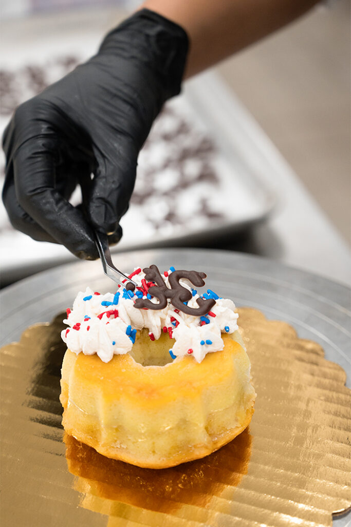 Side-by-side images show a chef’s gloved hands garnishing a Coconut Flancocho dessert. In the first image, colorful sprinkles are being added over whipped topping. In the second image, a chocolate Coquí frog decoration is being placed on top of the finished dessert.