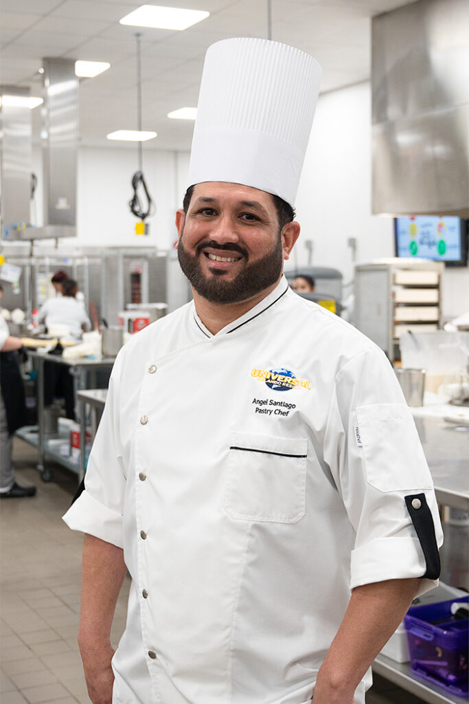 A Team Member in a white pastry chef uniform and tall chef hat stands in a commercial kitchen. Stainless steel equipment, workstations, and other staff are visible in the background.