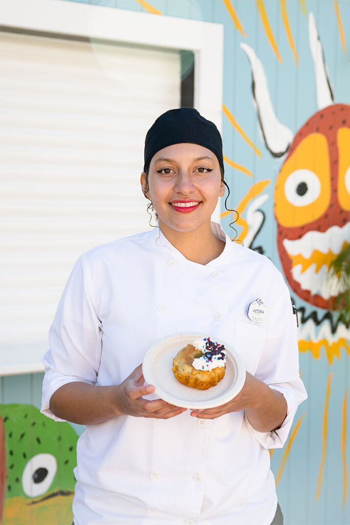 A Team Member wearing a white chef coat and black cap stands outdoors in front of a colorful mural, holding a plate with a Coconut Flancocho dessert topped with whipped cream, sprinkles and a chocolate Coquí frog decoration.