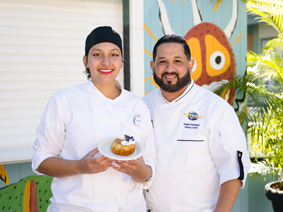Two Team Members wearing white chef uniforms stand outdoors in front of a colorful mural. One of them holds a plate featuring the Coconut Flancocho dessert from Universal Mardi Gras.