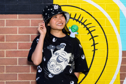 A woman stands against a brick and colorful wall wearing Minions and Monsters merch - bucket hat, t-shirt, and shoulder buddy