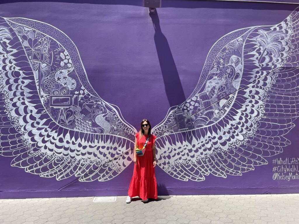 A woman in a pink dress stands in front of a purple mural with large white wings