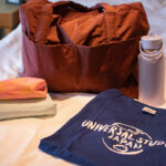 A bag and a handful of items (shirt, water bottle, smaller bags) sitting on a bed.