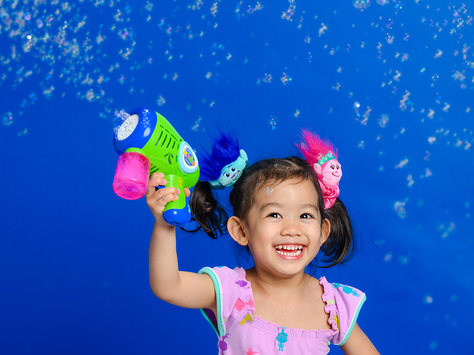 A little girl wearing a Trolls headband is blowing a bubble machine