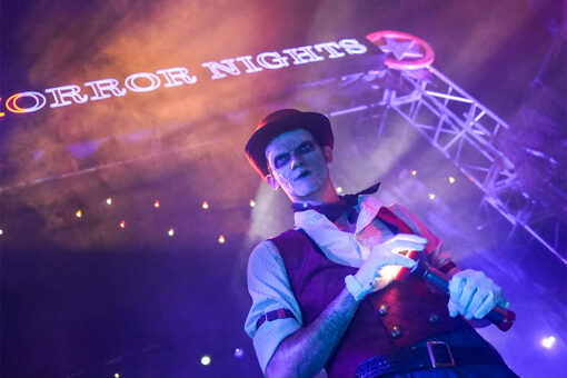 The Usher of HHN 2026 stands in front of the Halloween Horror Nights archway holding a flashlight.