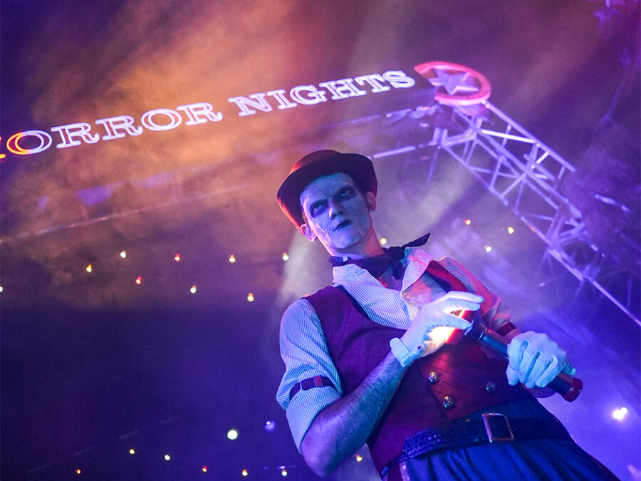 The Usher of HHN 2026 stands in front of the Halloween Horror Nights archway holding a flashlight.